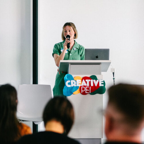 Alexis Bulman stands at a podium speaking into a microphone at the Creative PEI Arts, Access and Wellbeing conference. She wears a green outfit and speaks confidently to an audience seated in the foreground. A laptop rests on the podium, which features a colourful Creative PEI logo, in a bright, modern conference room.