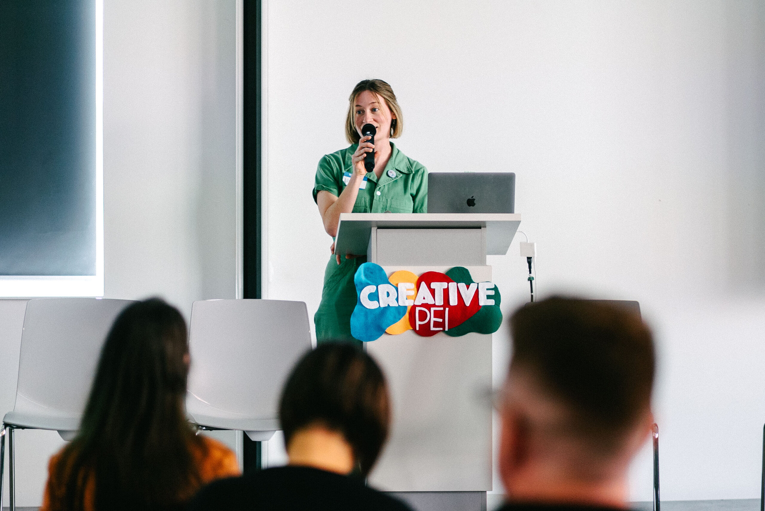 Alexis Bulman stands at a podium speaking into a microphone at the Creative PEI Arts, Access and Wellbeing conference. She wears a green outfit and speaks confidently to an audience seated in the foreground. A laptop rests on the podium, which features a colourful Creative PEI logo, in a bright, modern conference room.